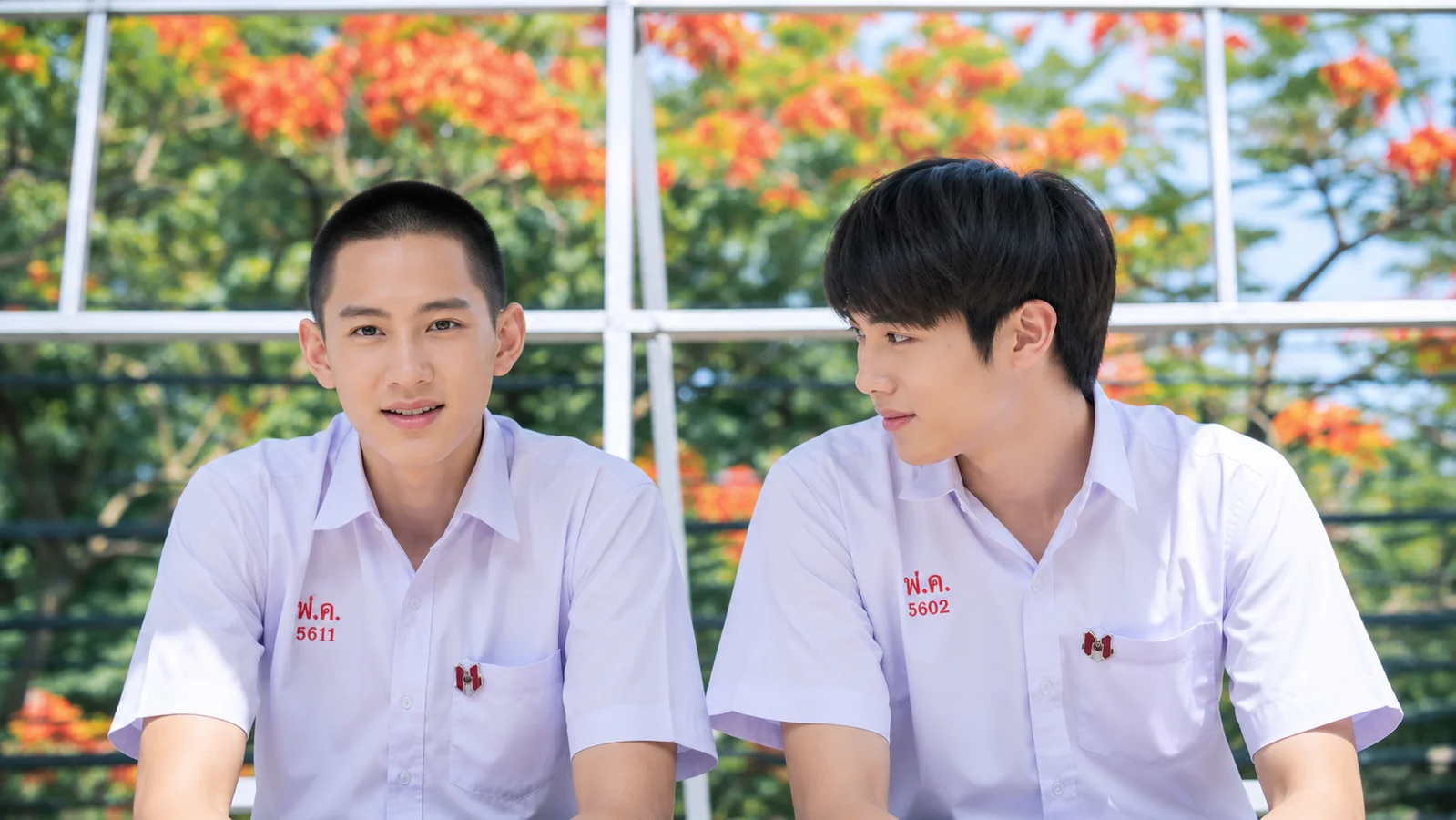 Punn and Noh from the 2014 Thai BL series Love Sick sit together in white school uniforms, with Noh looking toward Punn against a backdrop of green trees and orange flowers.