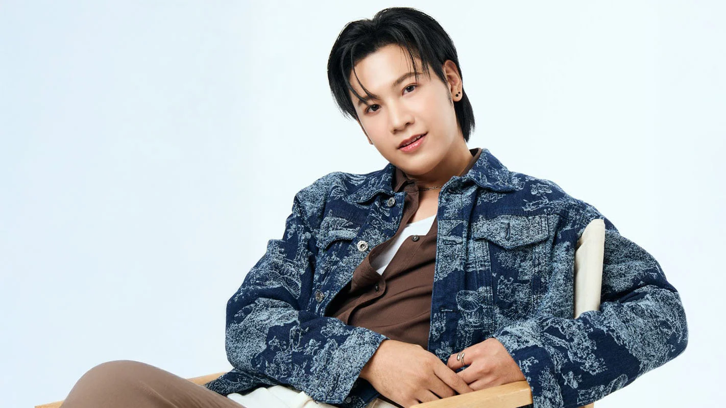 Singto Prachaya sits in a wooden chair wearing a blue patterned denim jacket against a pale blue background.