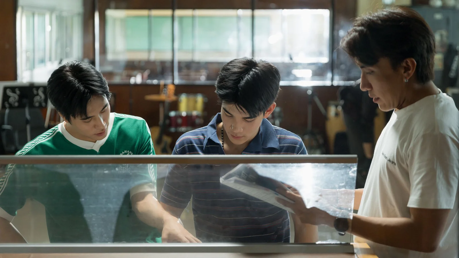 Gemini Norawit and Fourth Nattawat review materials at a piano while director P’Aof stands beside them holding an open binder.