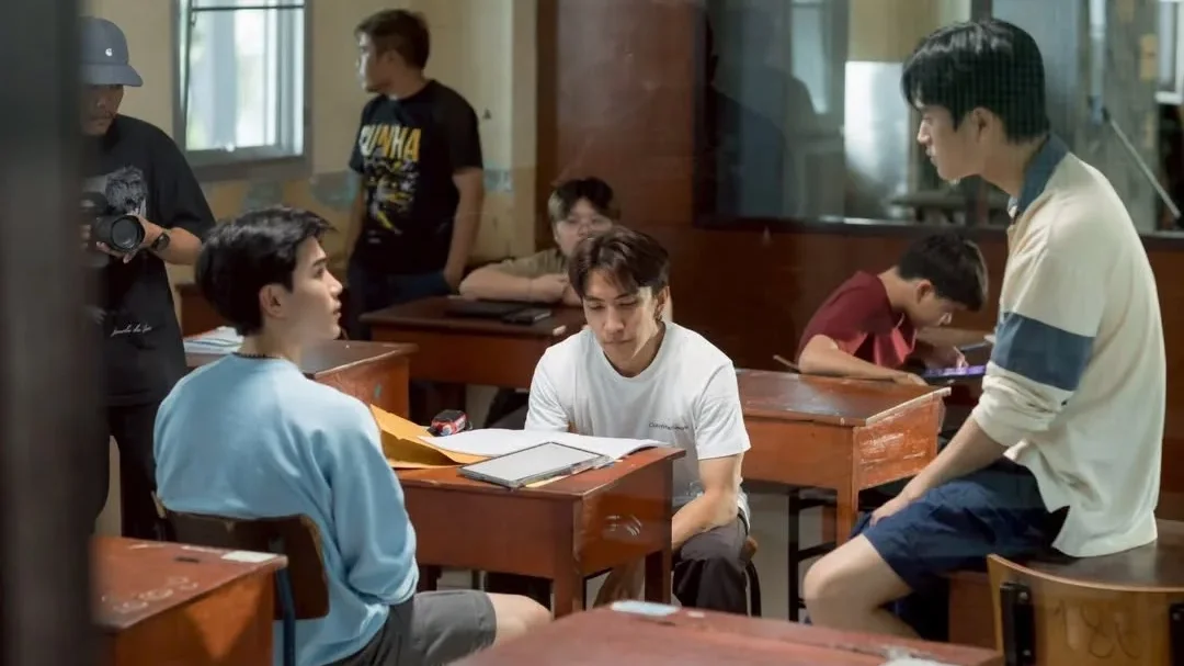 Gemini Norawit, Fourth Nattawat, and director P’Aof talk between takes on the Ticket to Heaven classroom set as crew members work around them.