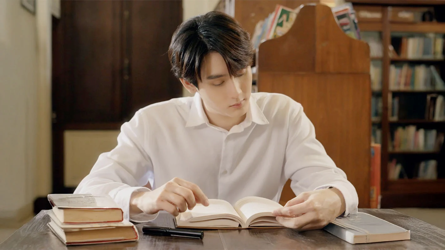 Book Kasidet Plookphol as Theo, seated at a library table reading a book in Enchanté.