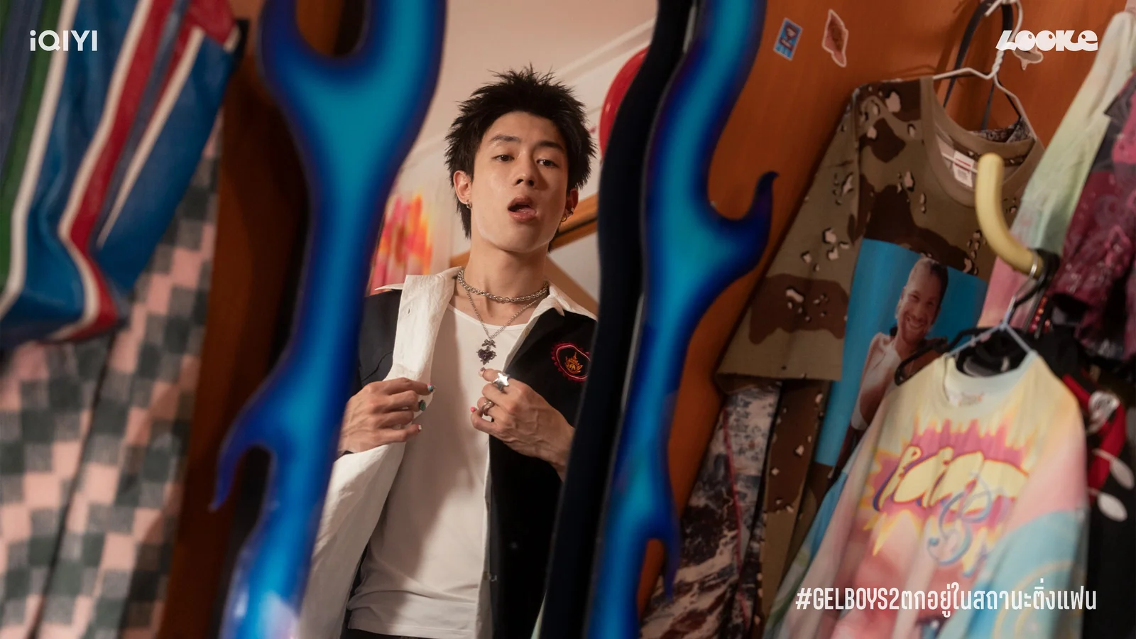 Fou4Mod, played by New Chayapak, adjusts his jacket between racks of colorful clothing.