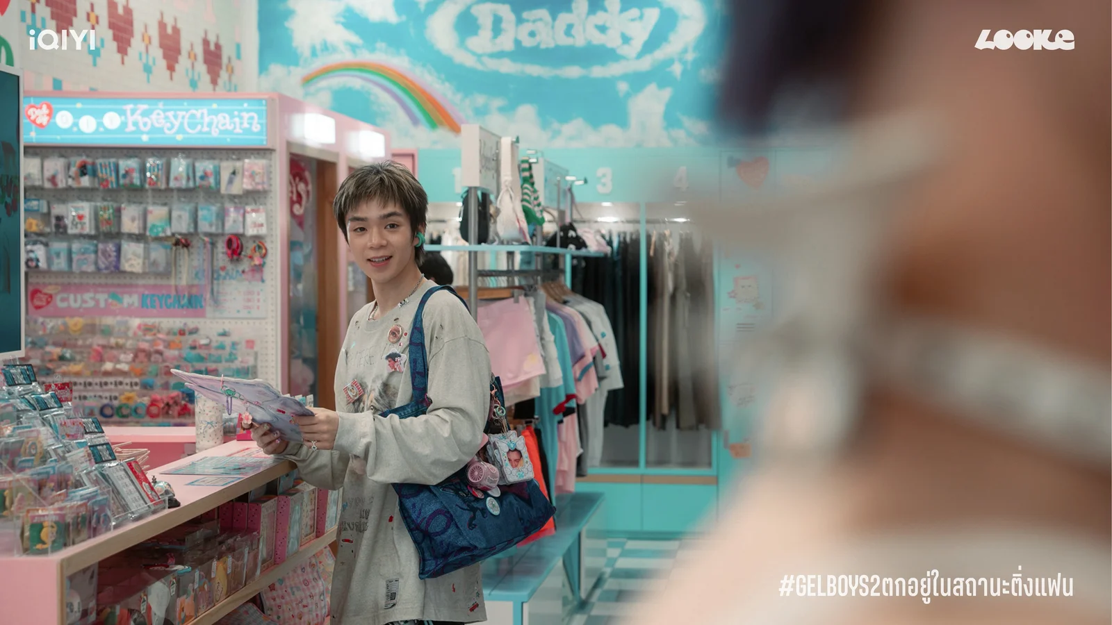 Baabin, played by PJ Mahidol, smiles while browsing a colorful keychain and accessories shop.