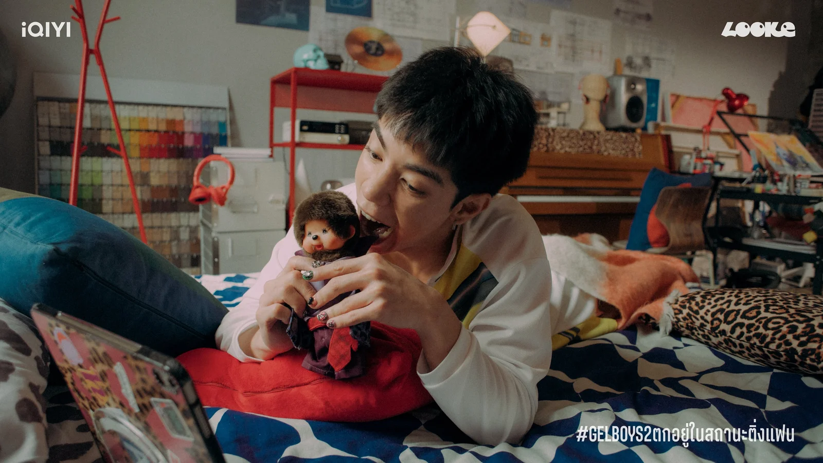 Chian, played by Pide Monthapoom, playfully bites a small doll while relaxing in a colorful bedroom.