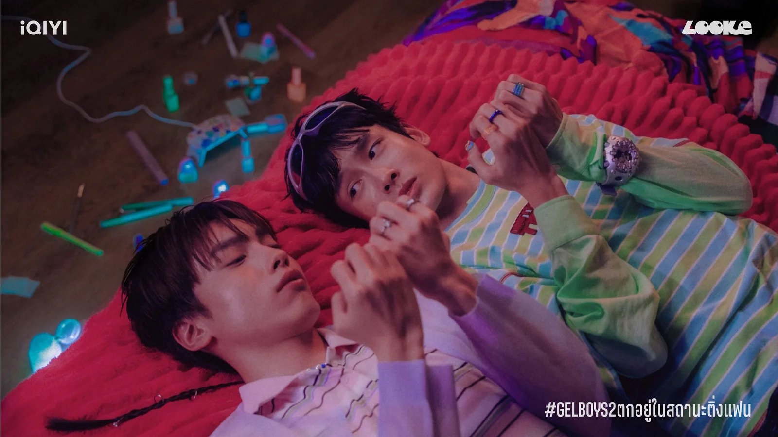 Gelz and Baby, played by Peem Veeravit and Mye Nattapat, lie together on a bright red rug while examining their painted nails.