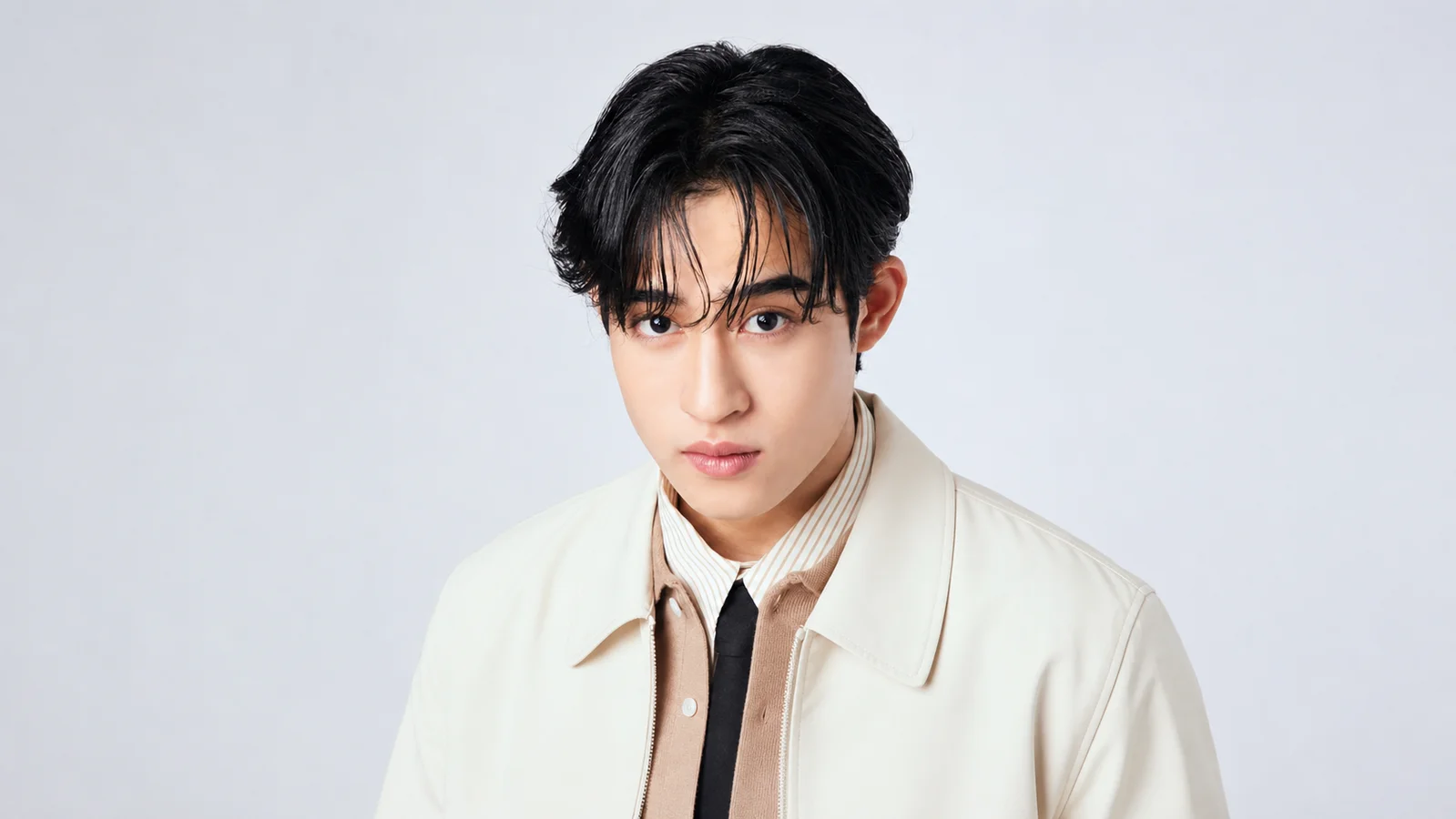 Barcode Tinnasit looks directly at the camera in a cream jacket, beige shirt and black tie against a pale gray studio background.