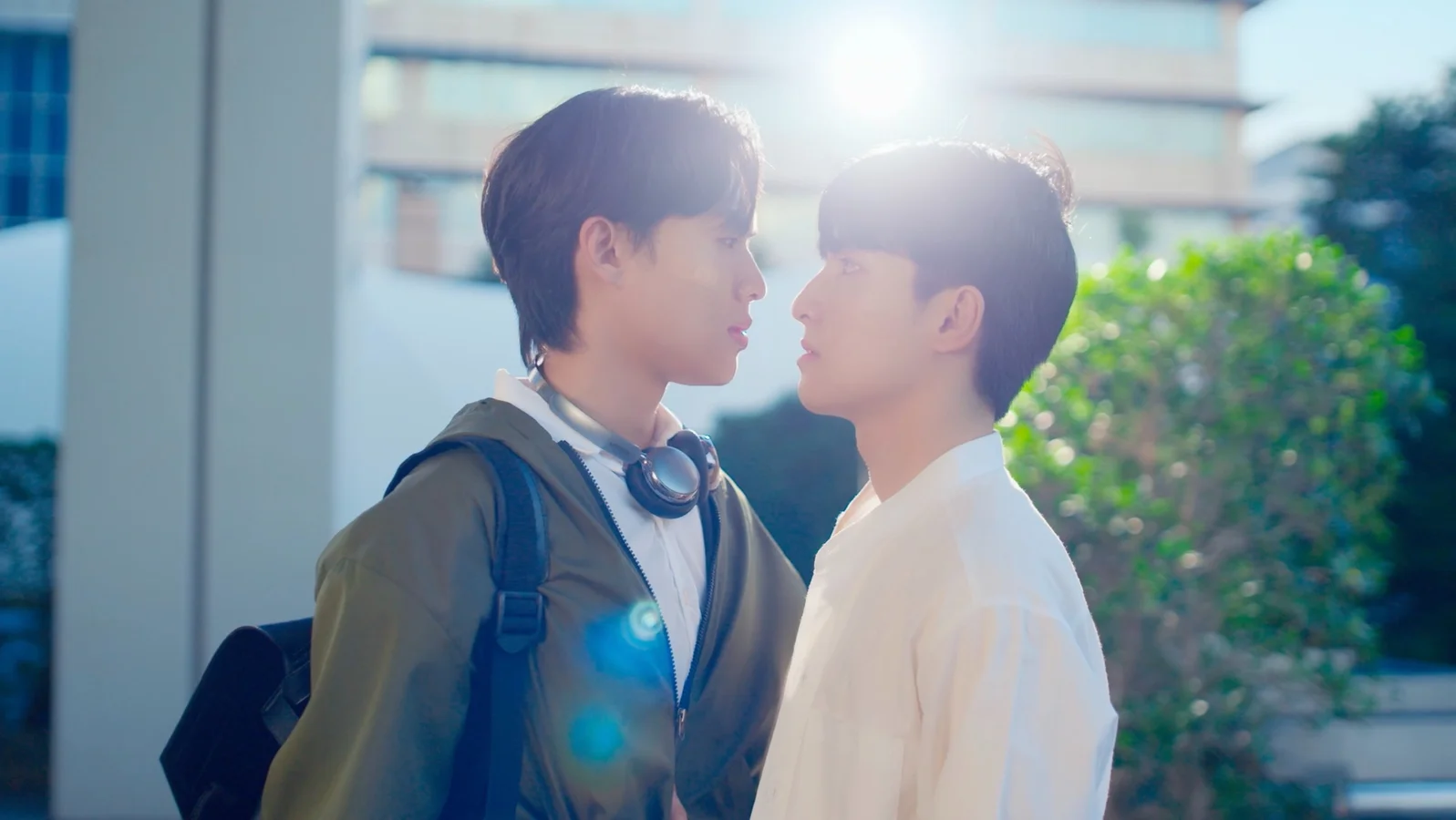 Rin, wearing headphones and a backpack, stands inches from Khobfah as they lock eyes in a brightly sunlit courtyard in Unlucky Bae.
