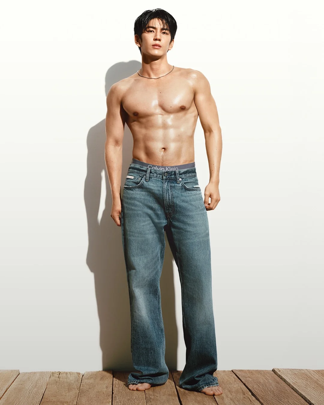 Sea Tawinan stands shirtless and barefoot in relaxed blue jeans against a sunlit white wall.