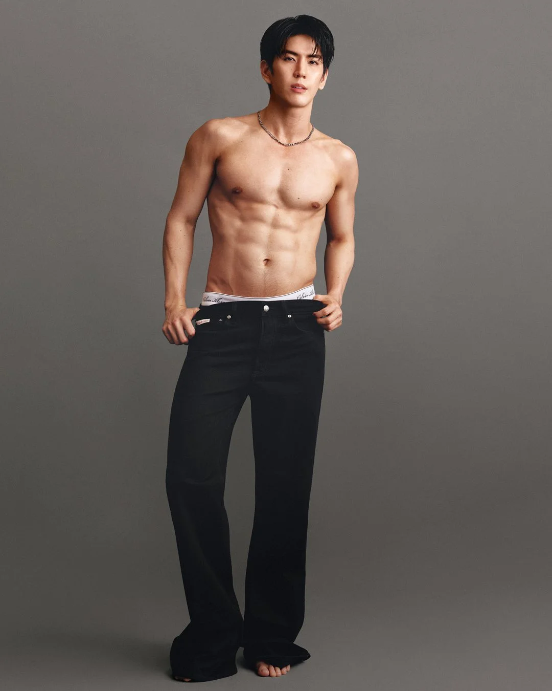 Sea Tawinan poses shirtless and barefoot in loose black jeans against a gray studio backdrop.