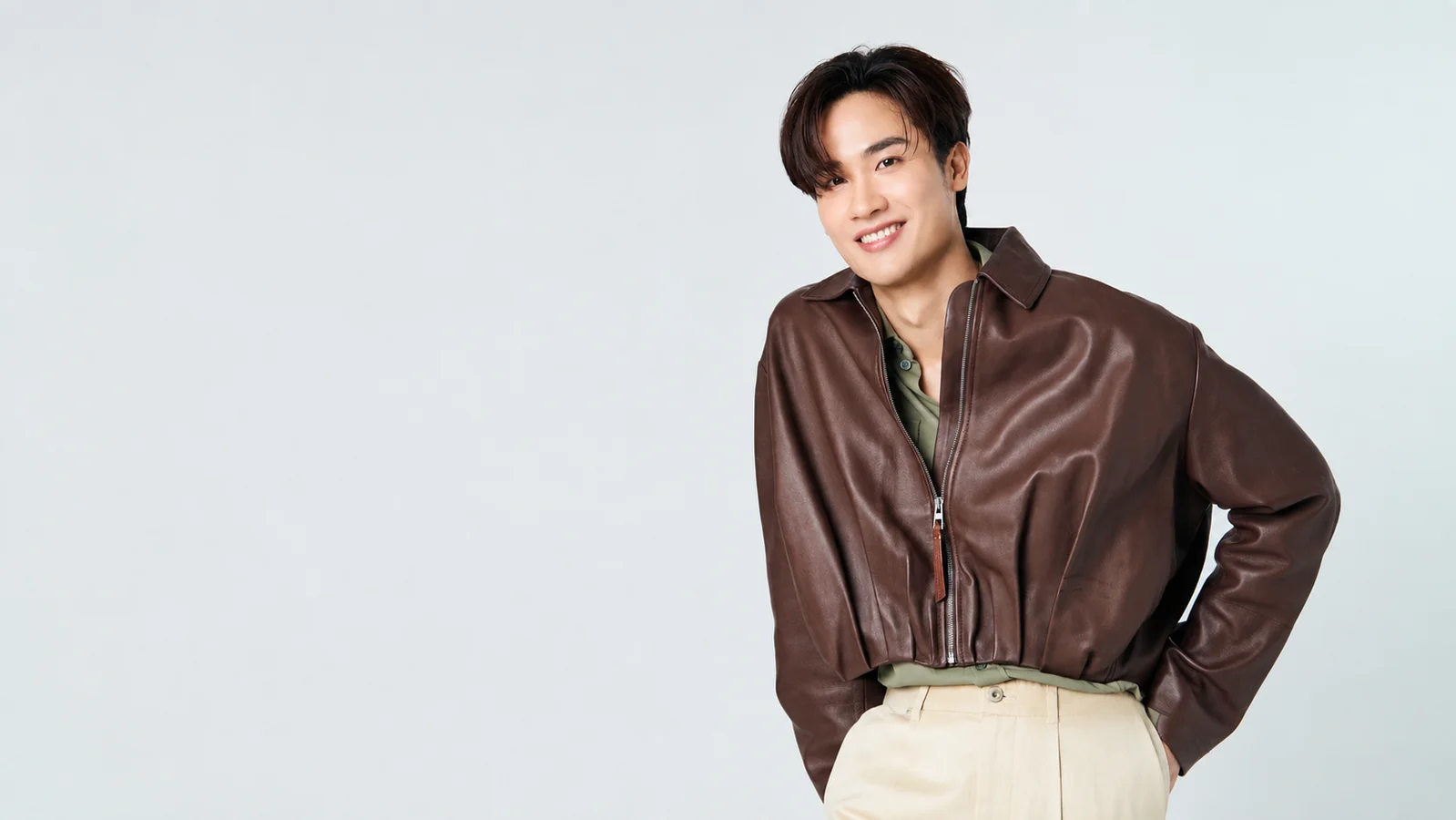 Tay Tawan smiles in a brown leather jacket, olive-green shirt and cream trousers against a pale studio background.
