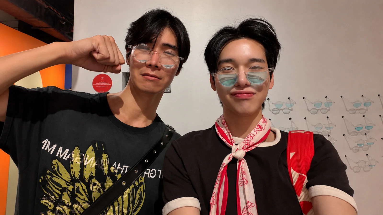 JT Patiphan and Ohm Thanakrit pose together in novelty glasses during an outing, with JT raising his fist on the left and Ohm wearing a red-and-white scarf on the right.