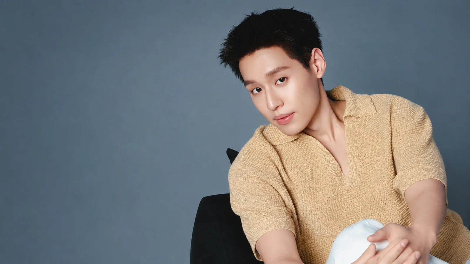 Billkin Putthipong poses in a beige knit shirt against a muted blue-gray studio background.