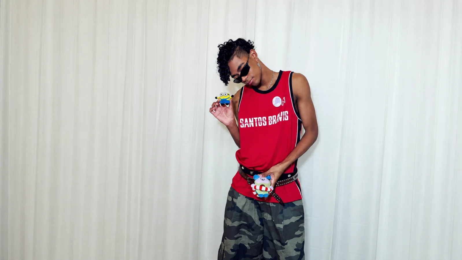 Kauê Penna of SANTOS BRAVOS poses in a red jersey and camouflage pants while holding a Minion figure and JimmySea’s AVOCEAN plush against a white curtain.