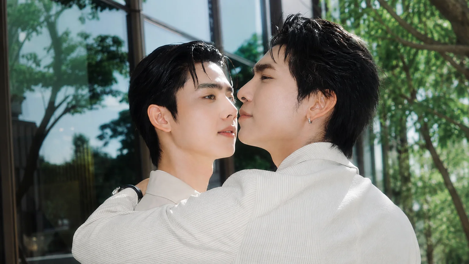 Mew Suppasit and Sunny Wannarat share a close embrace while posing together in coordinated light-colored outfits beside a glass building surrounded by greenery.