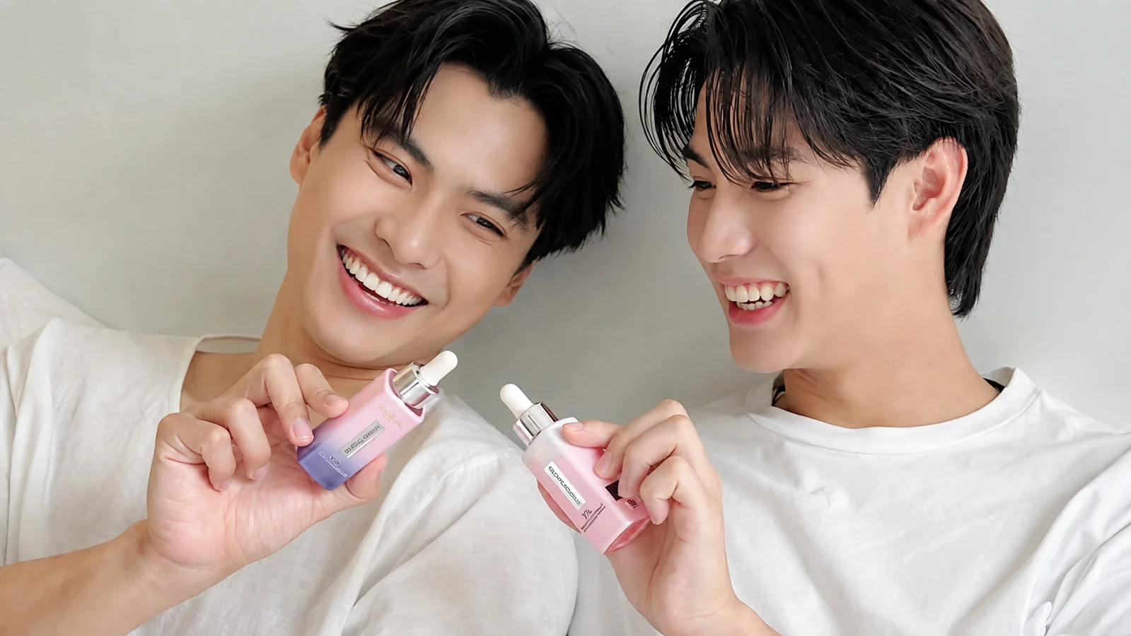 Ohm Pawat and Ing Asavanund smile beside each other in white shirts while holding pink L’Oréal Paris serum bottles during their first joint brand collaboration.